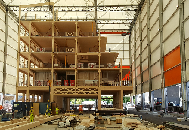 Construction of a multi-story wooden building inside a large industrial hall with workers and materials on the floor.