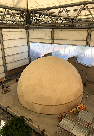 Large wooden dome structure under construction inside an industrial hall with steel beams and scaffolding.